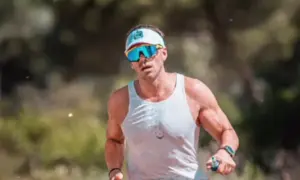 Hombre con gafas de sol deportivas y gorra, sudoroso, listo para salir a correr por un sendero boscoso.
