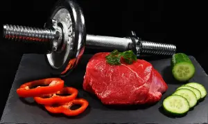 Un trozo de carne roja cruda, pimientos rojos y pepino junto a una pesa. La carne roja es clave para los beneficios del entrenamiento.