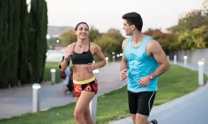 Pareja sonriente corre junta en un parque. Ella, con top deportivo y shorts coloridos, él, con camiseta azul. ¡Correr adelgaza y los hace felices!