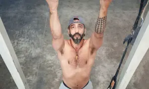 Hombre con barba y gorra realiza dominadas supinas en barra. Brazos fuertes, tatuaje geométrico en el brazo izquierdo.