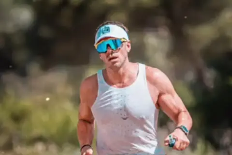Hombre con gafas de sol deportivas y gorra, sudoroso, listo para salir a correr por un sendero boscoso.