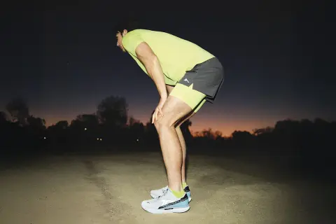 Corredor con las manos en las rodillas, recuperando el aliento al anochecer. Consejos para saber como evitar el flato al correr.