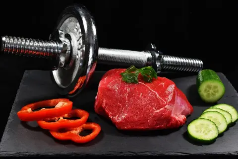 Un trozo de carne roja cruda, pimientos rojos y pepino junto a una pesa. La carne roja es clave para los beneficios del entrenamiento.
