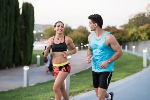 Pareja sonriente corre junta en un parque. Ella, con top deportivo y shorts coloridos, él, con camiseta azul. ¡Correr adelgaza y los hace felices!