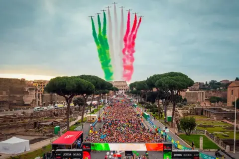 La **media maratón Roma** comienza con aviones dibujando la bandera italiana en el cielo, sobre una multitud de corredores y el Coliseo al fondo.