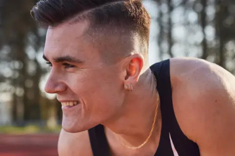 Un atleta sonríe, con su corte de pelo moderno y un collar dorado. Podría ser uno de los hermanos Ingebrigtsen, listo para competir.