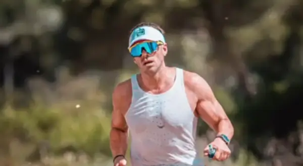 Hombre con gafas de sol deportivas y gorra, sudoroso, listo para salir a correr por un sendero boscoso.