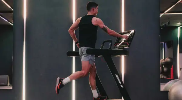 Hombre entrenando en cinta, ajustando la velocidad. El gimnasio moderno con luces LED crea un ambiente dinámico para el ejercicio.