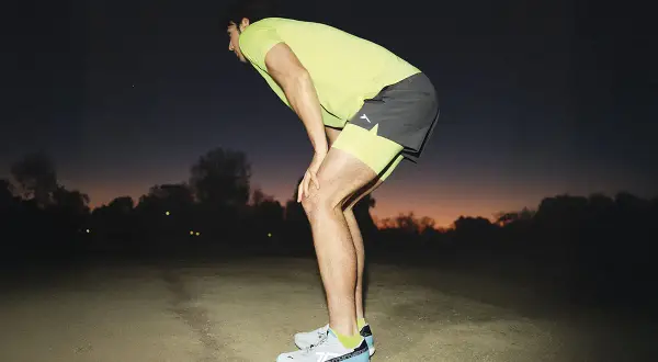 Corredor con las manos en las rodillas, recuperando el aliento al anochecer. Consejos para saber como evitar el flato al correr.