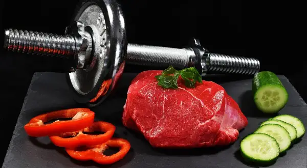 Un trozo de carne roja cruda, pimientos rojos y pepino junto a una pesa. La carne roja es clave para los beneficios del entrenamiento.