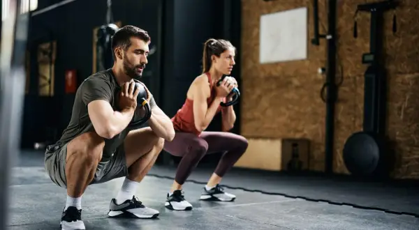 Pareja realiza sentadillas con kettlebell, parte de sus ejercicios para corredores, fortaleciendo piernas y core en el gimnasio.
