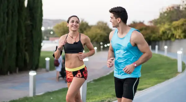 Pareja sonriente corre junta en un parque. Ella, con top deportivo y shorts coloridos, él, con camiseta azul. ¡Correr adelgaza y los hace felices!