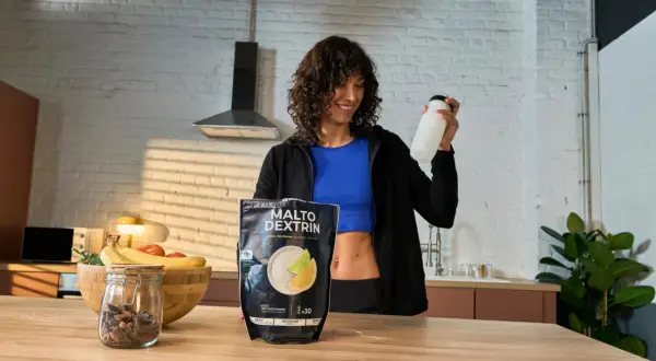 Mujer sonriente con una botella en la mano, junto a un paquete de MALTO DEXTRIN, un carbohidrato para energía.