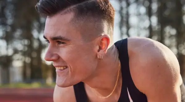 Un atleta sonríe, con su corte de pelo moderno y un collar dorado. Podría ser uno de los hermanos Ingebrigtsen, listo para competir.
