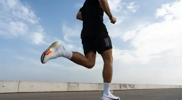 Un corredor con zapatillas blancas y naranjas practica como correr correctamente bajo un cielo azul con nubes.