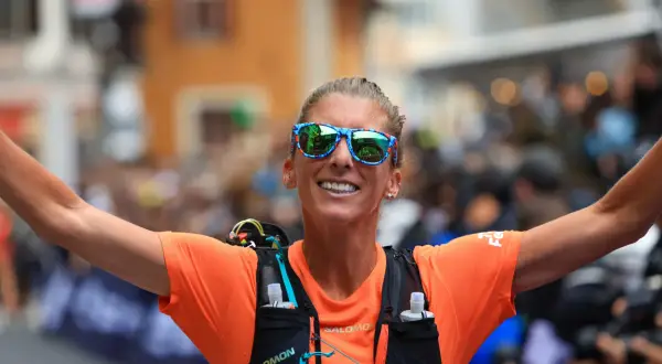 Courtney Dauwalter, con gafas de sol llamativas y camiseta naranja, celebra con los brazos extendidos tras una carrera.
