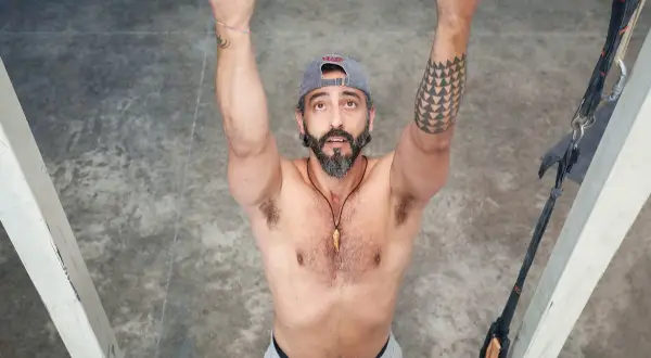 Hombre con barba y gorra realiza dominadas supinas en barra. Brazos fuertes, tatuaje geométrico en el brazo izquierdo.