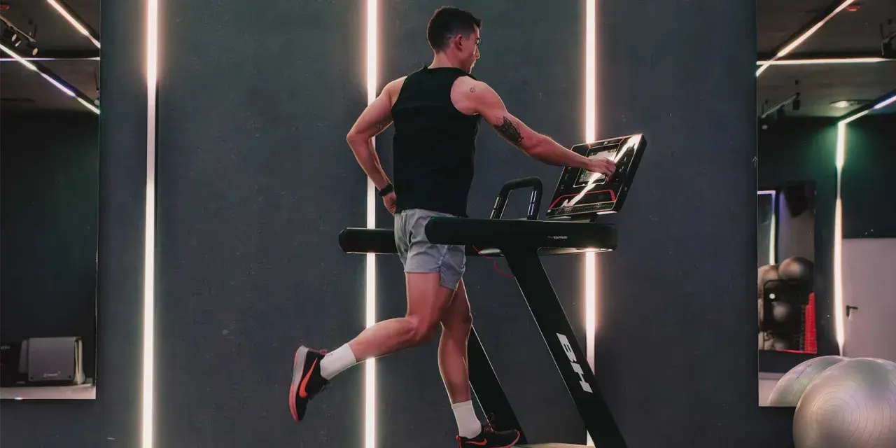 Hombre entrenando en cinta, ajustando la velocidad. El gimnasio moderno con luces LED crea un ambiente dinámico para el ejercicio.