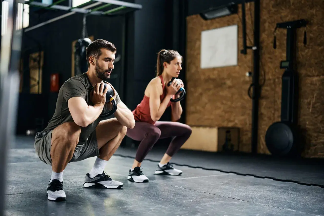 Pareja realiza sentadillas con kettlebell, parte de sus ejercicios para corredores, fortaleciendo piernas y core en el gimnasio.