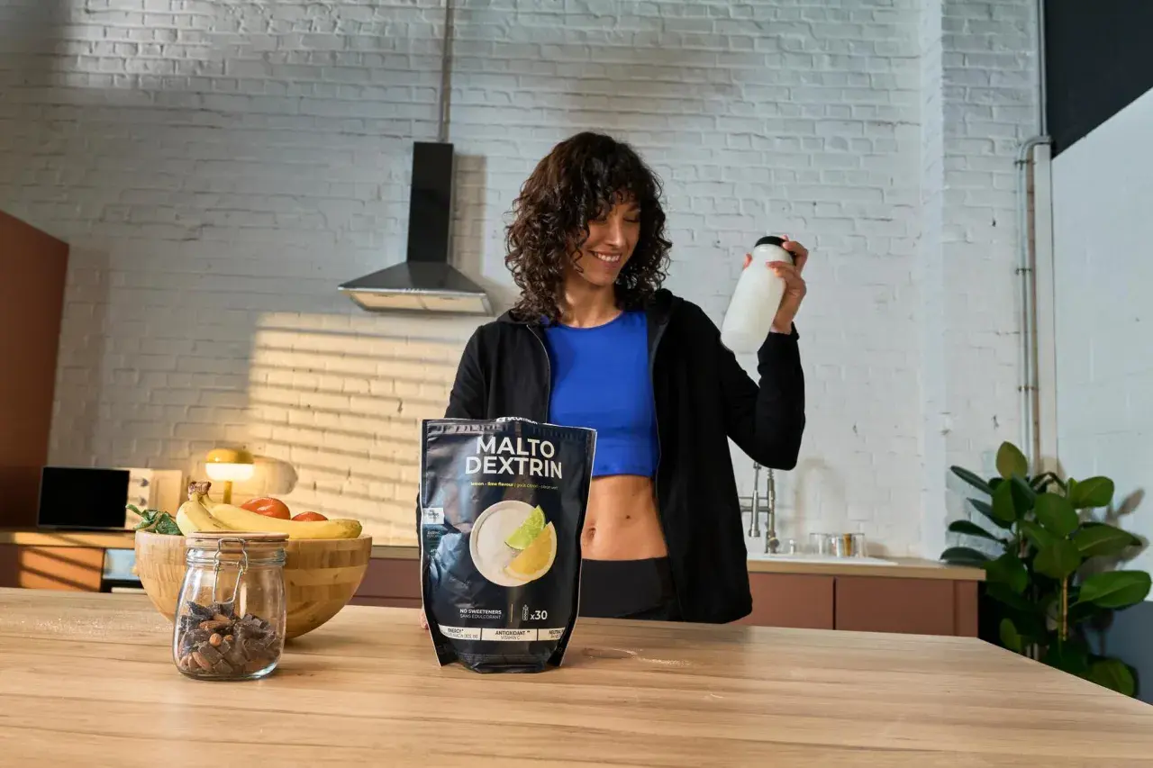 Mujer sonriente con una botella en la mano, junto a un paquete de MALTO DEXTRIN, un carbohidrato para energía.