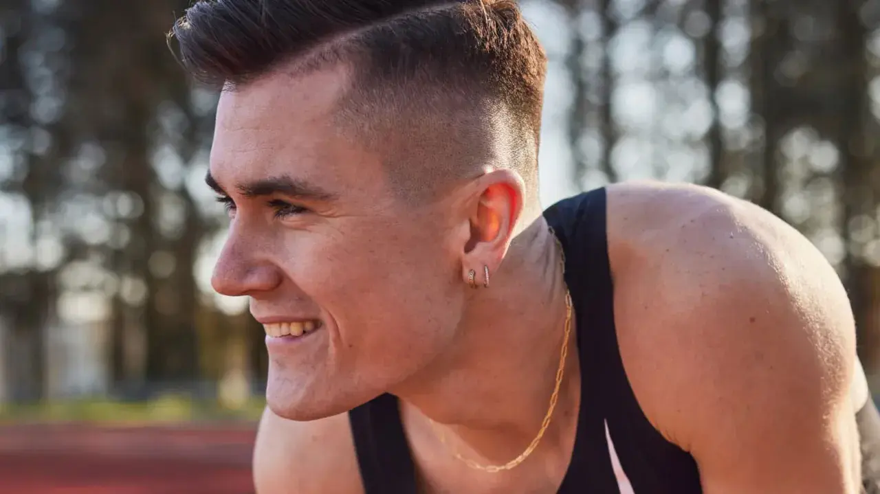 Un atleta sonríe, con su corte de pelo moderno y un collar dorado. Podría ser uno de los hermanos Ingebrigtsen, listo para competir.