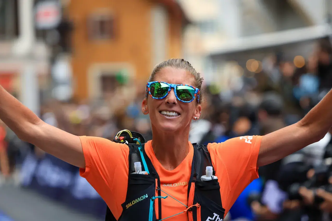 Courtney Dauwalter, con gafas de sol llamativas y camiseta naranja, celebra con los brazos extendidos tras una carrera.