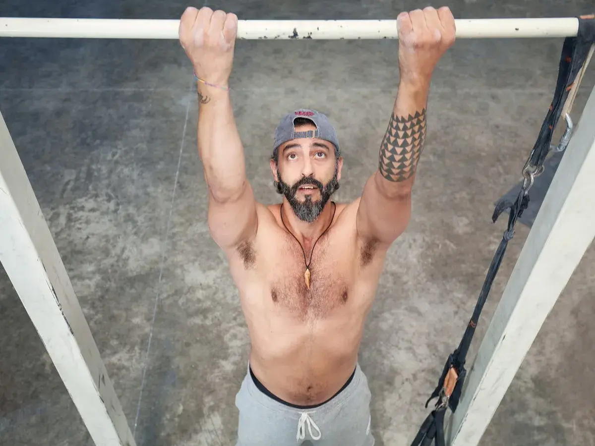 Hombre con barba y gorra realiza dominadas supinas en barra. Brazos fuertes, tatuaje geométrico en el brazo izquierdo.