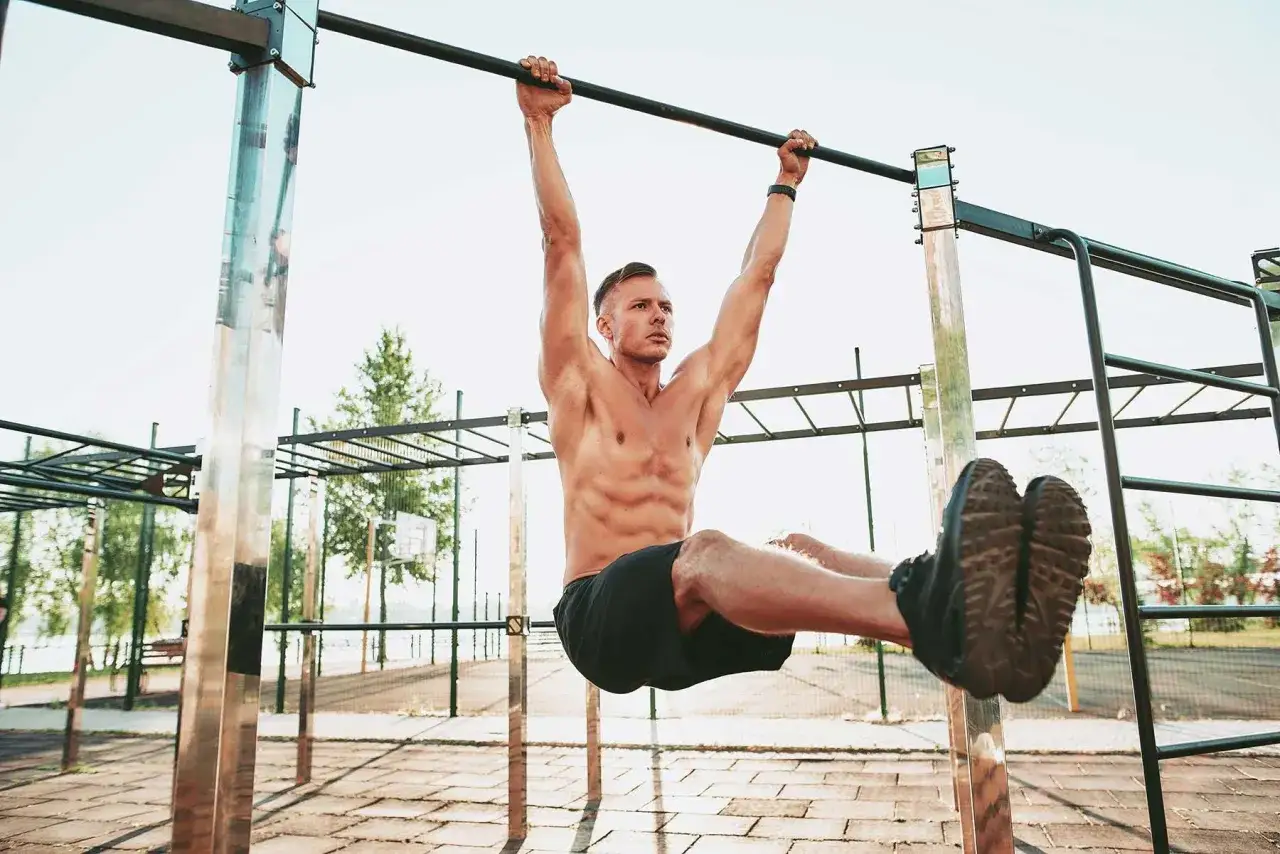 Hombre musculoso realizando elevación de piernas en barra, parte de su entrenamiento calistenia al aire libre.