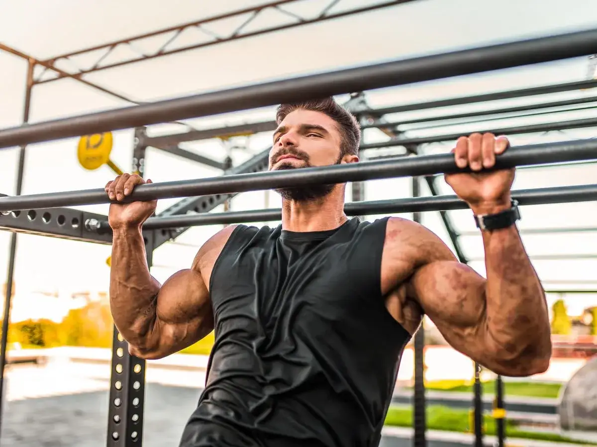 Hombre musculoso entrenando cómo hacer dominadas en una barra exterior.