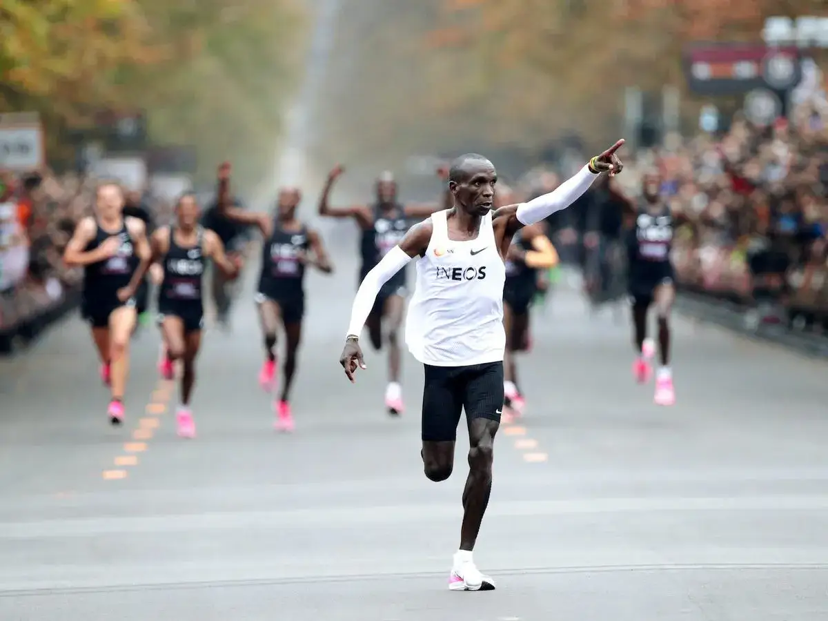 Eliud Kipchoge lidera la carrera, señalando hacia adelante. Otros corredores le siguen de cerca en el maratón.