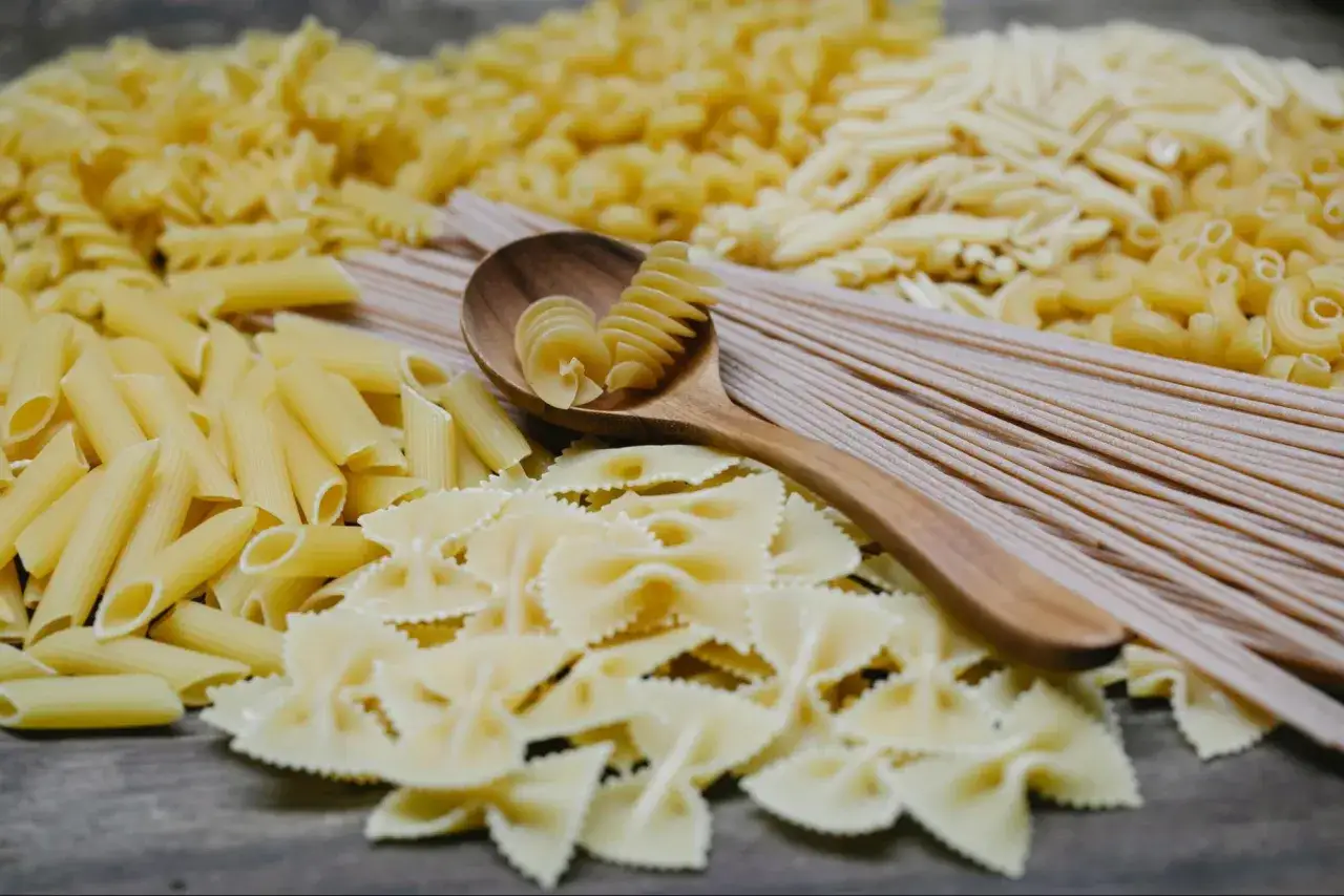 Variedad de pastas secas, incluyendo penne, farfalle y espagueti. Una cuchara de madera sostiene fusilli, mostrando la amilopectina que es un carbohidrato complejo.