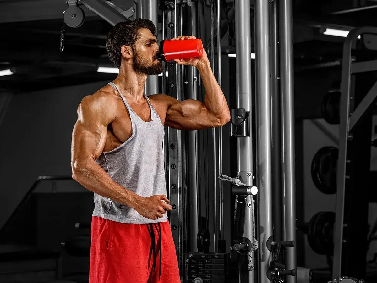 Hombre musculoso bebe proteína de un shaker rojo en el gimnasio, aprendiendo como tomar proteina para sus músculos.