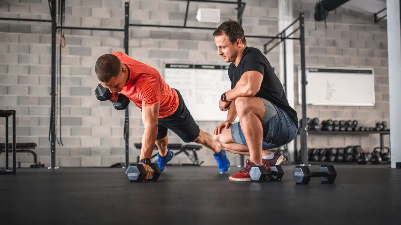 Un hombre realiza un remo con mancuerna en posici&oacute;n de plancha, mientras un personal trainer que es su gu&iacute;a, observa atentamente.