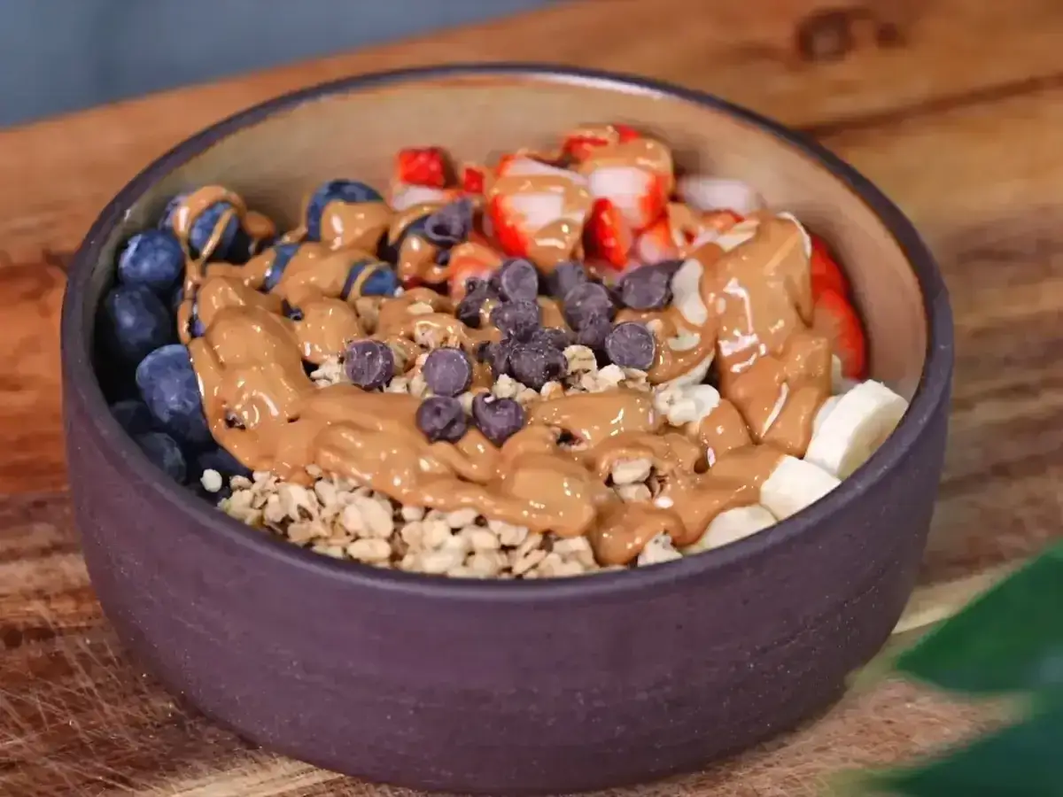 Un tazón lleno de arándanos, fresas, plátano y granola, cubierto con mantequilla de maní y chispas de chocolate. ¡La opción perfecta para que picar entre horas saludable!