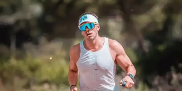 Hombre con gafas de sol deportivas y gorra, sudoroso, listo para salir a correr por un sendero boscoso.