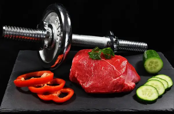 Un trozo de carne roja cruda, pimientos rojos y pepino junto a una pesa. La carne roja es clave para los beneficios del entrenamiento.