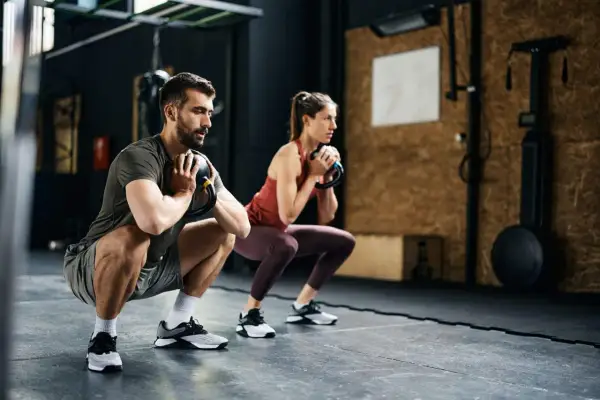 Pareja realiza sentadillas con kettlebell, parte de sus ejercicios para corredores, fortaleciendo piernas y core en el gimnasio.