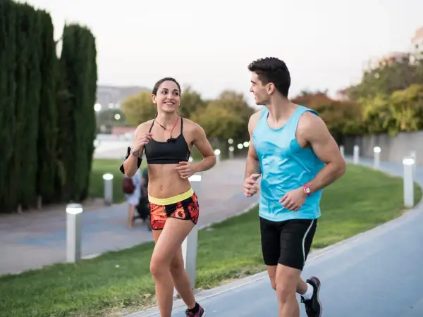 Pareja sonriente corre junta en un parque. Ella, con top deportivo y shorts coloridos, él, con camiseta azul. ¡Correr adelgaza y los hace felices!