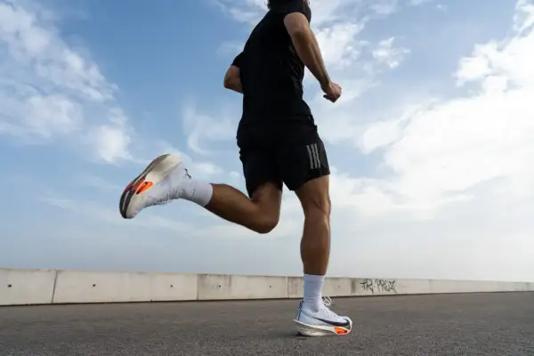 Un corredor con zapatillas blancas y naranjas practica como correr correctamente bajo un cielo azul con nubes.