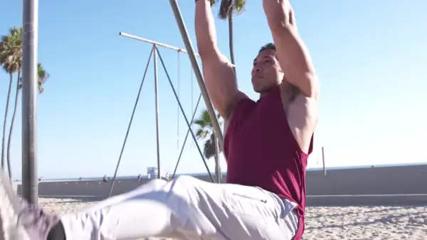 Hombre haciendo abdominales en barra en la playa, con palmeras y el mar de fondo.
