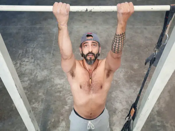 Hombre con barba y gorra realiza dominadas supinas en barra. Brazos fuertes, tatuaje geométrico en el brazo izquierdo.
